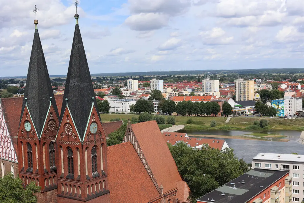 Kirchtürme ragen über Dächer und Fluss. Architektur mit roten Ziegeln und Turmspitzen. Stadtbild im Hintergrund.